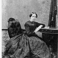 Image: woman in long dress sitting on a chair and leaning on a table. 