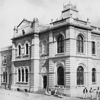 Image: An imposing, two-storey stone building in Victorian-Italianate style stands at the intersection of two dirt streets. A small group of men and boys stand in front of the building