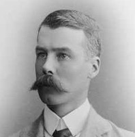Image: A photographic head-and-shoulders portrait of a young Caucasian man wearing a light-coloured, late-Victorian era suit and sporting a very large handlebar moustache