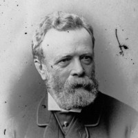 Image: A photographic head-and-shoulders portrait of a bearded, middle-aged man wearing a suit with cravat and jewelled cravat pin
