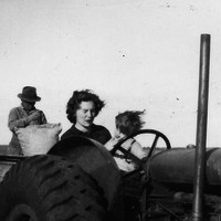A woman and a small child sitting behind the steering wheel of a tractor.  A man behind them is sewing up a hessian bag.