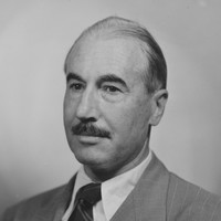 Image: A formal photographic head-and-shoulders portrait of a balding, moustachioed middle-aged man in a suit and tie
