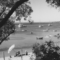 Image: Black and white photograph of people on a beach, with boats in the water in the distance.