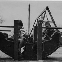 Image: Two boys sit in a ‘rocking boat’ at a playground. A swings-set is positioned immediately behind the boys