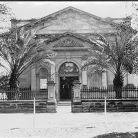 Image: Black and white photograph of the front of a building. Palm trees stand on either side of the front of the building. The building reads "ART GALLERY"