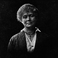 Image: Black and white photograph of woman wearing a dark jacket over a white blouse with a cross around her neck. Her light coloured hair is either pinned back or cut short.