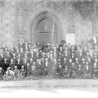 Image: Black and white group photograph of over 150 people. There are five rows of people, mostly men, standing in front of a church