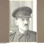 Image: A moustachioed middle-aged Caucasian man in an Australian Army officer's uniform. He is wearing a hat and spectacles with oval lenses