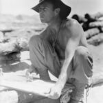 Image: A man crouching down holding a drawing board, and a pencil and looking into the distance