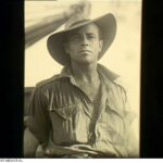 Image: Upper body portrait photograph of a man wearing a shirt with sleeves rolled up, and a large wide brimmed hat