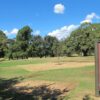 Image: a grassy park with a variety of species of trees. In the foreground is a  red and green sign with the name of the park (Rymill Park/Mullawirraburka), some other printed text which cannot be made out in this photograph, and a map.