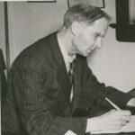 Image: A middle-aged, clean-shaven Caucasian man in a 1950s vintage pin-striped suit sits at a desk and writes on a piece of paper. He appears to be deep in thought as he writes