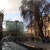 Image: A modern, glass-fronted building connects two multi-storey historic buildings. A modern fountain sculpture is in the foreground