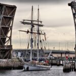 Image: A two-masted sailing vessel passes through a drawbridge on a cloudy morning. Another bridge crossed by modern auto-mobiles is visible in the background