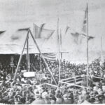 Image: a crowd of people in 1870s clothing gather around a small stage where a large foundation stone is being lowered into place.