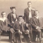 Image: Four men and a woman in university graduation attire sit on chairs and pose for a photograph