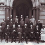 Image: A group of men in Edwardian attire pose for a photograph. A single woman in a light-coloured outfit is seated among their ranks in the front row