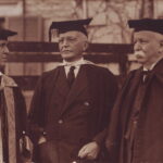 Image: Three men dressed in suits and full academic regalia speak to one another outside a building. Two of the men are middle-aged senior academics, while the other is younger and appears to be in a junior academic role