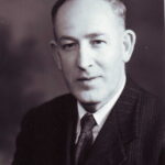 Image: A photographic head-and-shoulders portrait of a clean-shaven, middle-aged Caucasian man wearing a suit of 1940s vintage