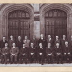 Image: A group of several middle-aged and elderly Caucasian men and two elderly Caucasian women pose for a photograph in front of a large stone building. The group is wearing business attire of 1950s vintage