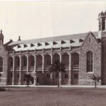 Image: the side view of a large stone hall with towers, windows set within stone arches and a pitched roof. A lawn with palm trees and a statue of a man can be seen in the foreground.