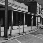 Image: A line of historic buildings line an asphalt street. Many of the buildings appear to be either abandoned or in a neglected state