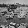 Image: A bridge under construction extends across a river in an active port. Several sail-boats and other small watercraft are moored within a basin in the foreground