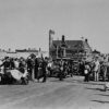 Image: A procession of 1940s-era cars escorted by a policeman on a motorcycle travels through a crowd of people gathered on the approach to a bridge