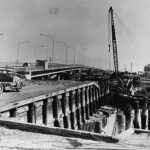 Image: Large cranes dismantle an old bridge span over a narrow river. Cars drive across a new bridge located a short distance and parallel to the old one