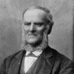 Image: A photographic portrait of a middle-aged balding man in Victorian attire. He is sporting a long chin-beard