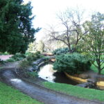 Image: A creek winds through a landscaped wooded parkland. A footpath follows the course of the creek