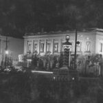 Image: A large plinth at the intersection of two streets with the words ‘Keep Left’ painted on it is illuminated at night. Buildings in the background are also illuminated, including one with the word ‘Centenary’ spelled out in its upper floor windows