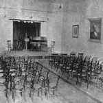 Image: A large, open hall with a stage at one end. Several rows of chairs face the stage, which features additional chairs, a music stand, and a piano