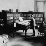 Image: A small group of men in a book-filled library read from desks