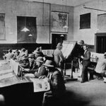 Image: A group of men in hats sit at two long tables and read newspapers