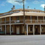 Image: Colour photograph of a two-storey late Victorian-era building on the corner of two paved streets. The building is painted tan with white columns and has the words ‘Railway Hotel, 1849’ painted beneath its roof