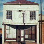 Image: A two-storey building with brick first floor and corrugated metal second floor stands derelict next to a city street