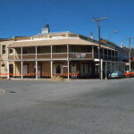 Image: Colour photograph of a two-storey late Victorian-era building on the corner of two paved streets. The building is painted tan with white columns and has the words ‘Railway Hotel, 1849’ painted beneath its roof