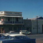 Image: A cream coloured two-storey building with second-floor verandah. Two cars are parked in front of the building and a man stands near its front door