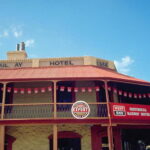 Image: Colour photograph of a two-storey late Victorian-era building on the corner of two paved streets. The building is painted tan with red trim and has the words ‘Rail ay Hotel, 1856’ painted beneath its roof