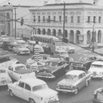 Image: A throng of auto-mobiles of 1940s, 1950s and 1960s vintage circle around a large plinth with the words ‘Keep Left’ painted on it