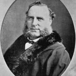 Image: A photographic head-and-shoulders portrait of a Caucasian man with large mutton-chop sideburns wearing late Victorian-era clothing, including an overcoat with fur collar