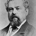 Image: A photographic head-and-shoulders portrait of a middle-aged Caucasian man with curly salt-and-pepper hair and goatee. He is wearing a late nineteenth-century suit and tie