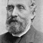 Image: A photographic head-and-shoulders portrait of a bearded middle-aged man in a tweed suit with tie