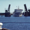 Image: A large, modern steel-hulled ferry passes through an open drawbridge. Part of a wharf is visible in the foreground