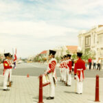 A group of men wearing red soldiers uniforms stand in two lines facing in front of a wide street extending into distance