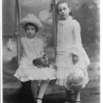 Image: Two young girls pose for a portrait. One sits on a swing, the other stands beside her. They are wearing white, frilly dresses.