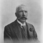 Image: Portrait photograph of a man wearing a suit and tie. He has a large mustache and his hair is white