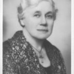 Image: Portrait photograph of a woman in her late fifties/early sixties with short light hair. She is wearing a cross necklace.