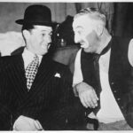 Image:Smiling man in suit and hat next to man in make-up and dirty vest